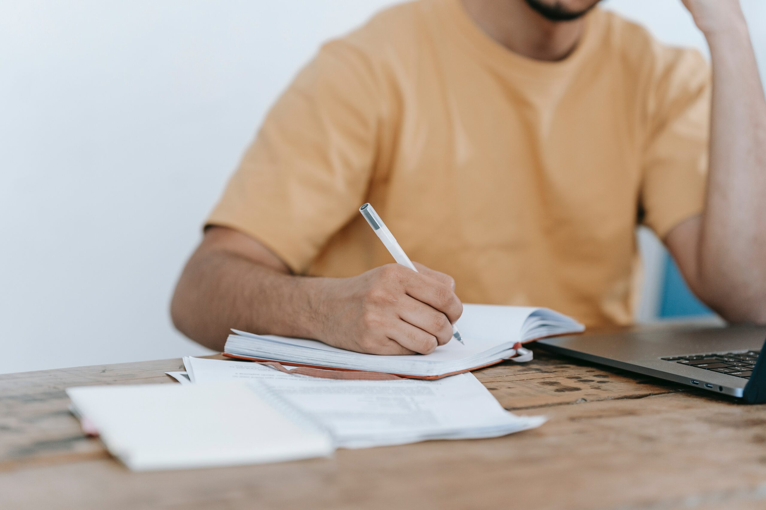 A person writing in a notebook with a laptop open next to him.